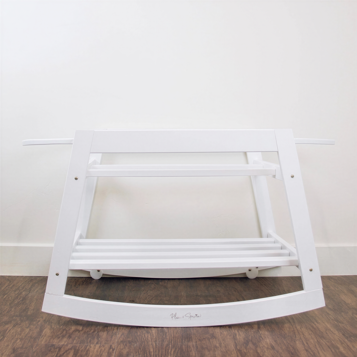 A white wooden rocking chair with a shelf underneath, placed on a wooden floor against a white wall.