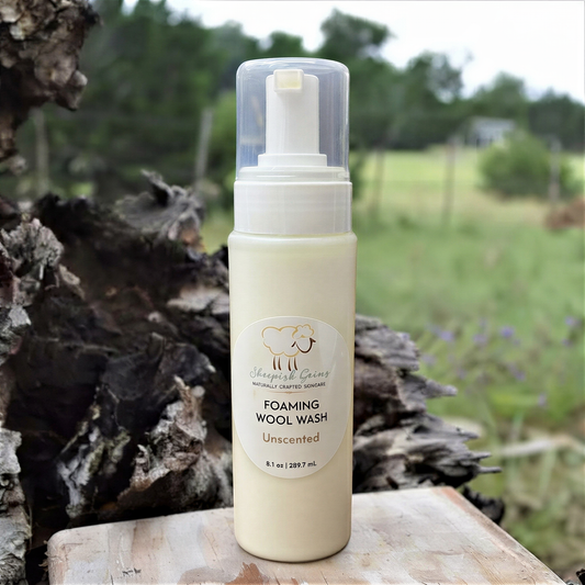 A bottle of Sheepish Gains Foaming Wool Wash Unscented, with a label that reads "Unscented" and "8 fl oz (1.39 fl oz)". The bottle is placed on a wooden surface, with a blurred background of trees and grass.