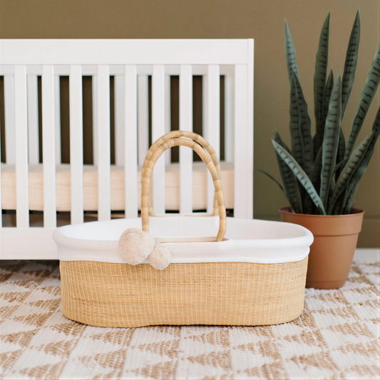 A beige woven basket with a handle sits on a patterned rug in a nursery setting, accompanied by a potted plant and a white crib.