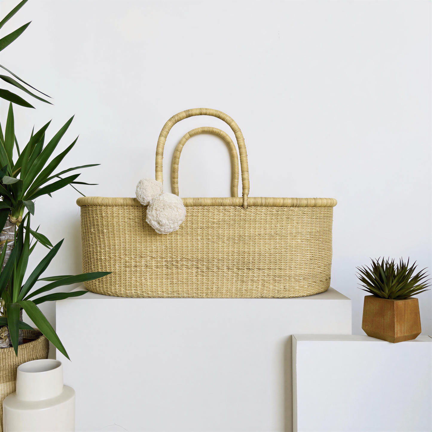 a woven basket with a handle, placed on a white surface, accompanied by a potted plant and a wooden box.